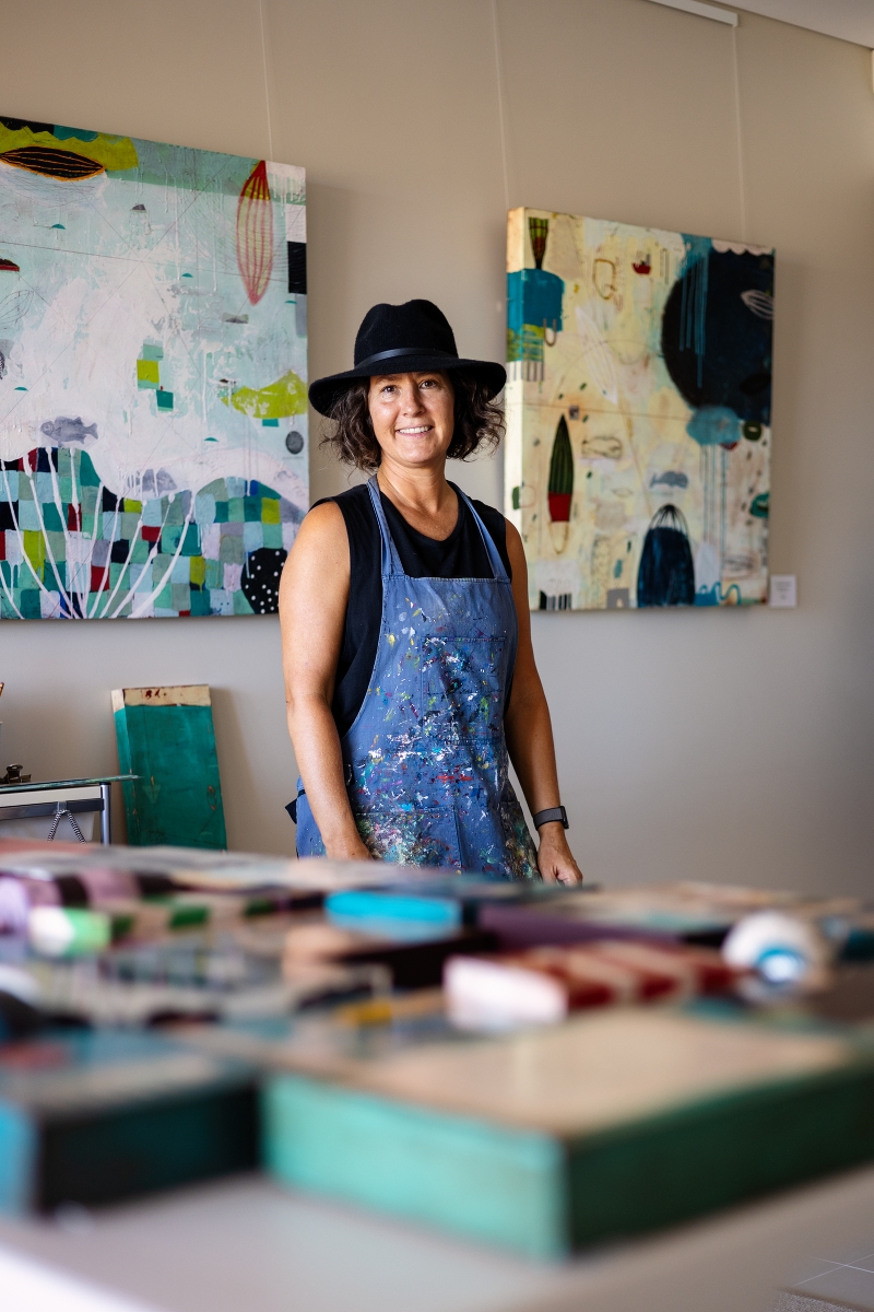 Artist standing in a studio with abstract paintings displayed on the wall.